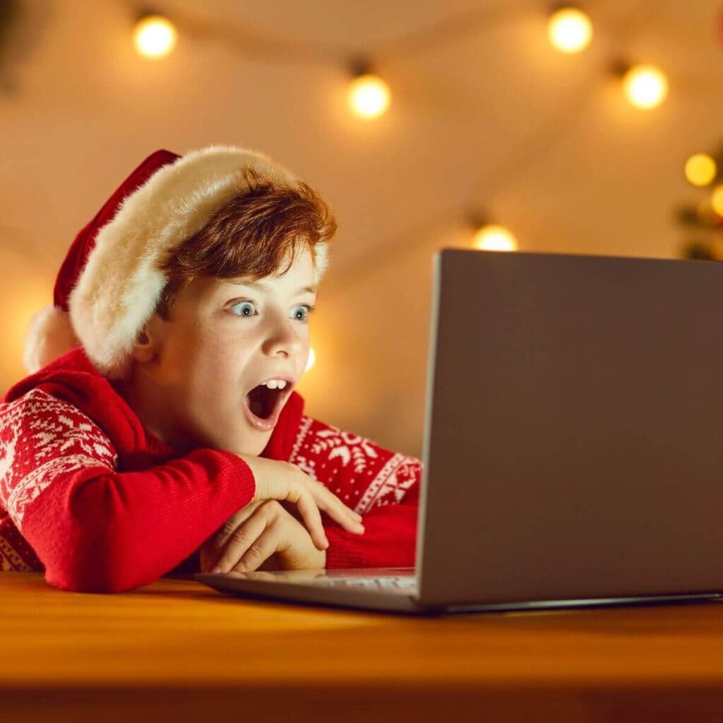 A boy wearing a Santa hat and a red sweater is sitting at a table looking at a laptop screen with his mouth open in shock. There are Christmas lights on the wall behind him.