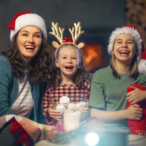 A woman wearing a Santa had and blue sweater is sitting on a couch with a girl wearing reindeer antlers and another girl who is wearing a Santa hat. They are all smiling.