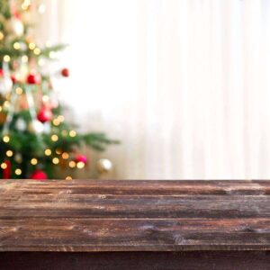 A Christmas tree is in front of a window and there is a wooden table at the front of the picture.