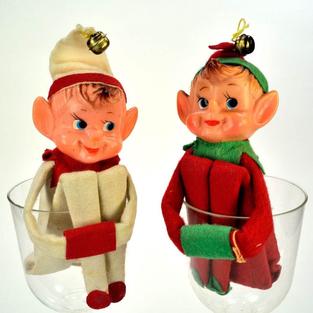 One elf wearing a cream colored suit with red trim is sitting in a clear plastic cup. Another elf is sitting in a clear plastic cup and his outfit is red with green trim