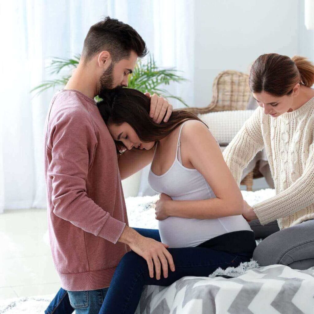 A pregnant woman is sitting on the side of the bed. A man is sitting in front of her and a woman is rubbing the lower part of her back.