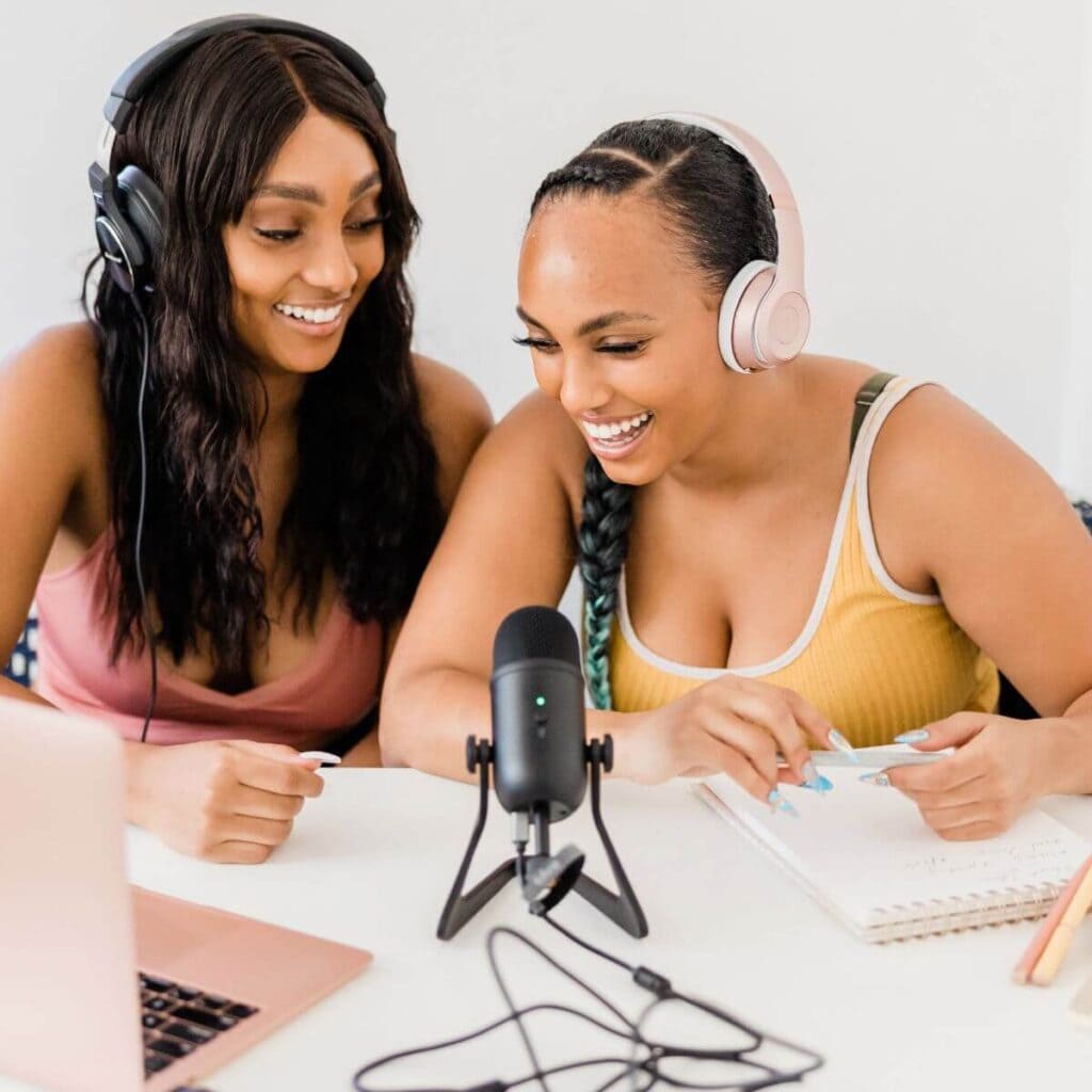 Two African American women are sitting at a white table. They have a pink laptop and black microphone in front of them. They are both wearing headphones and smiling.