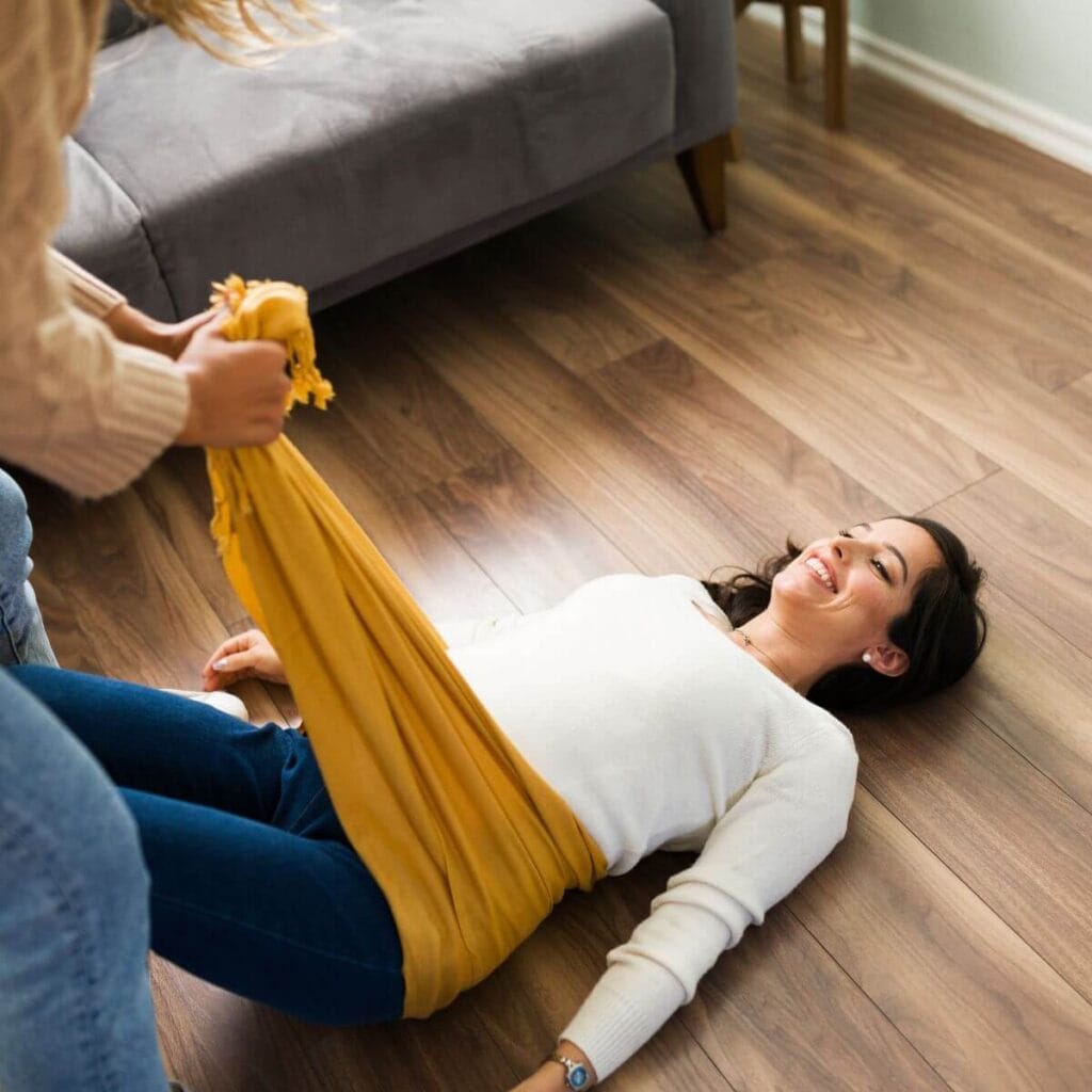 A pregnant woman in a white shirt and jeans is lying on the floor with a mustard yellow blanket around her hips while a woman pulls the blanket up