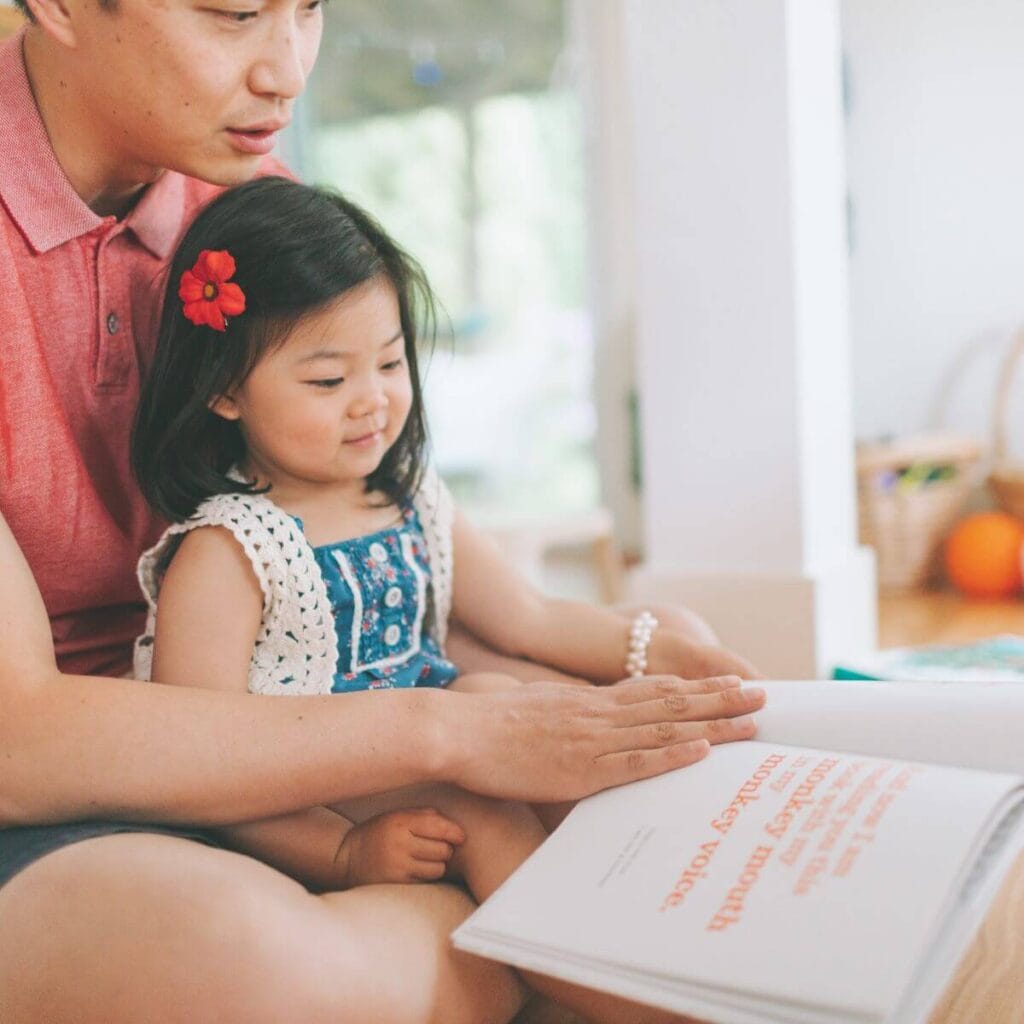 An Asian American dad is sitting on a wooden floor. His daughter is sitting in his lap and they are looking at a page in the book he is holding.