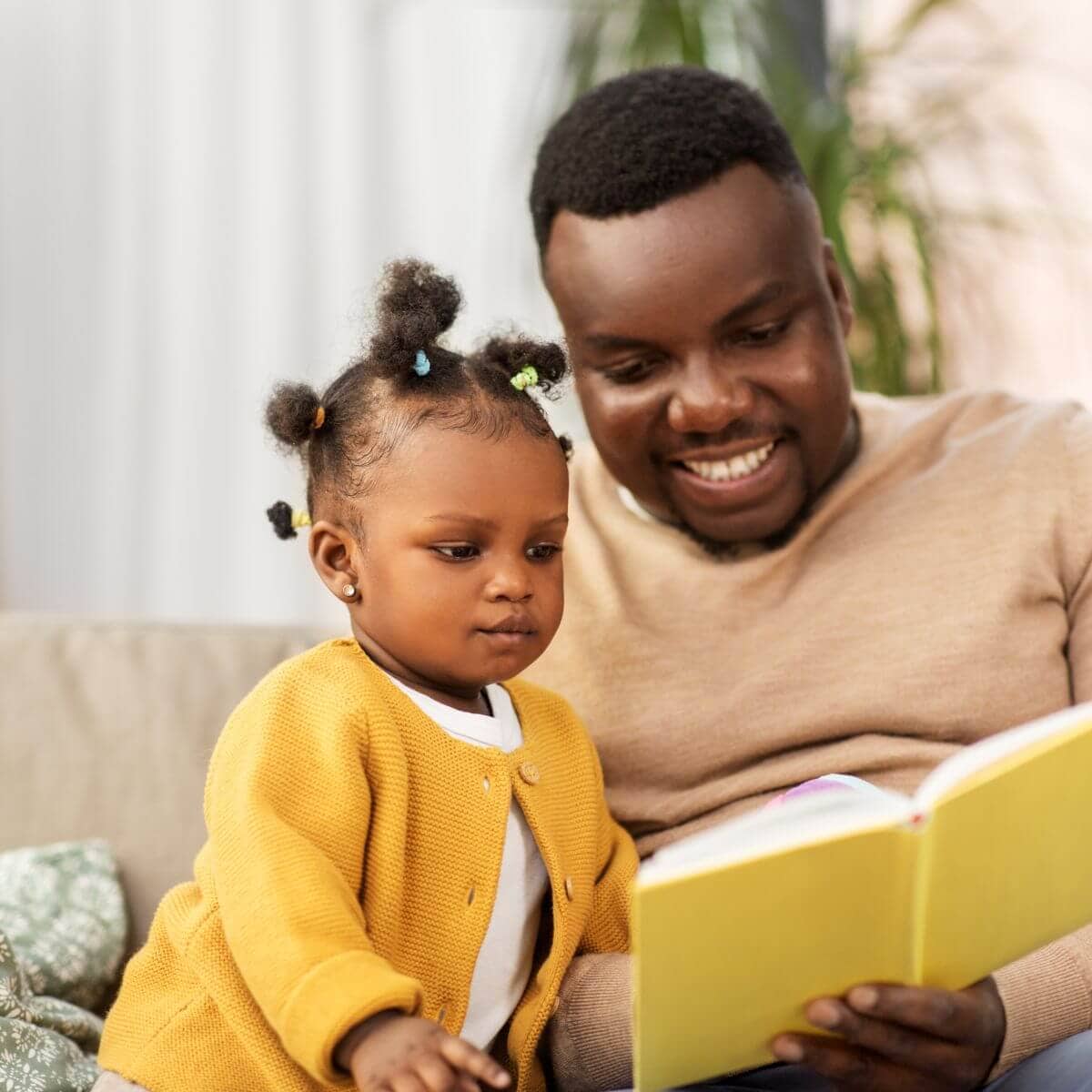 An African American dad is sitting down with his young daughter on a couch. He is holding a light yellow book and they are both looking at it.