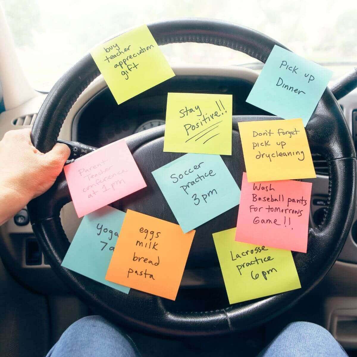 A woman is sitting in a vehicle holding on to the steering wheel. On the steering wheel are yellow, blue, pink, and orange post it notes with everything she needs to do.