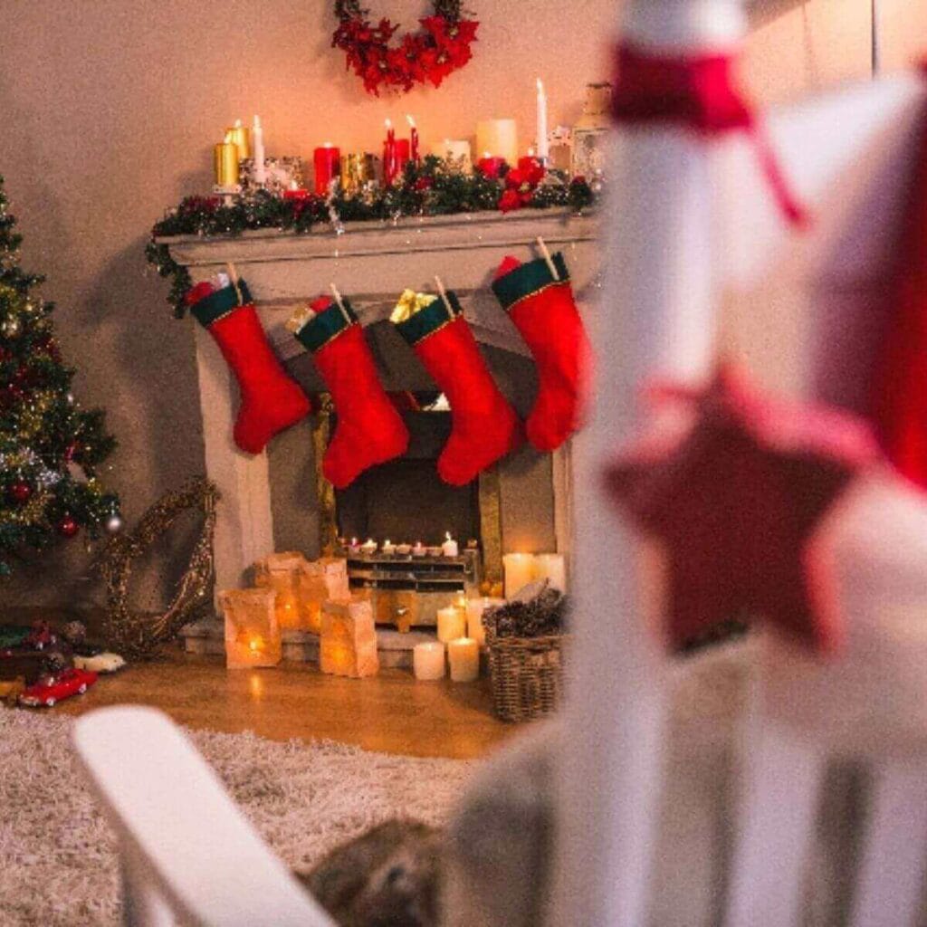 A white rocking chair is sitting in a living room. There is a decorated Christmas tree in the left hand corner of the room. In the middle is a fireplace with four stockings hanging from it.