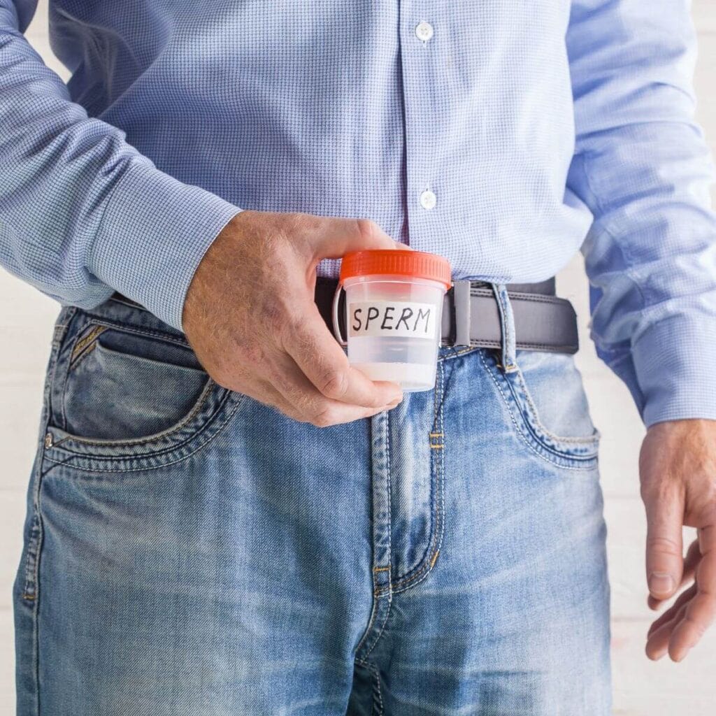 A man wearing a blue and white checkered shirt and light colored blue jeans is holding a clear cup with an orange top in his hand that says "Sperm."