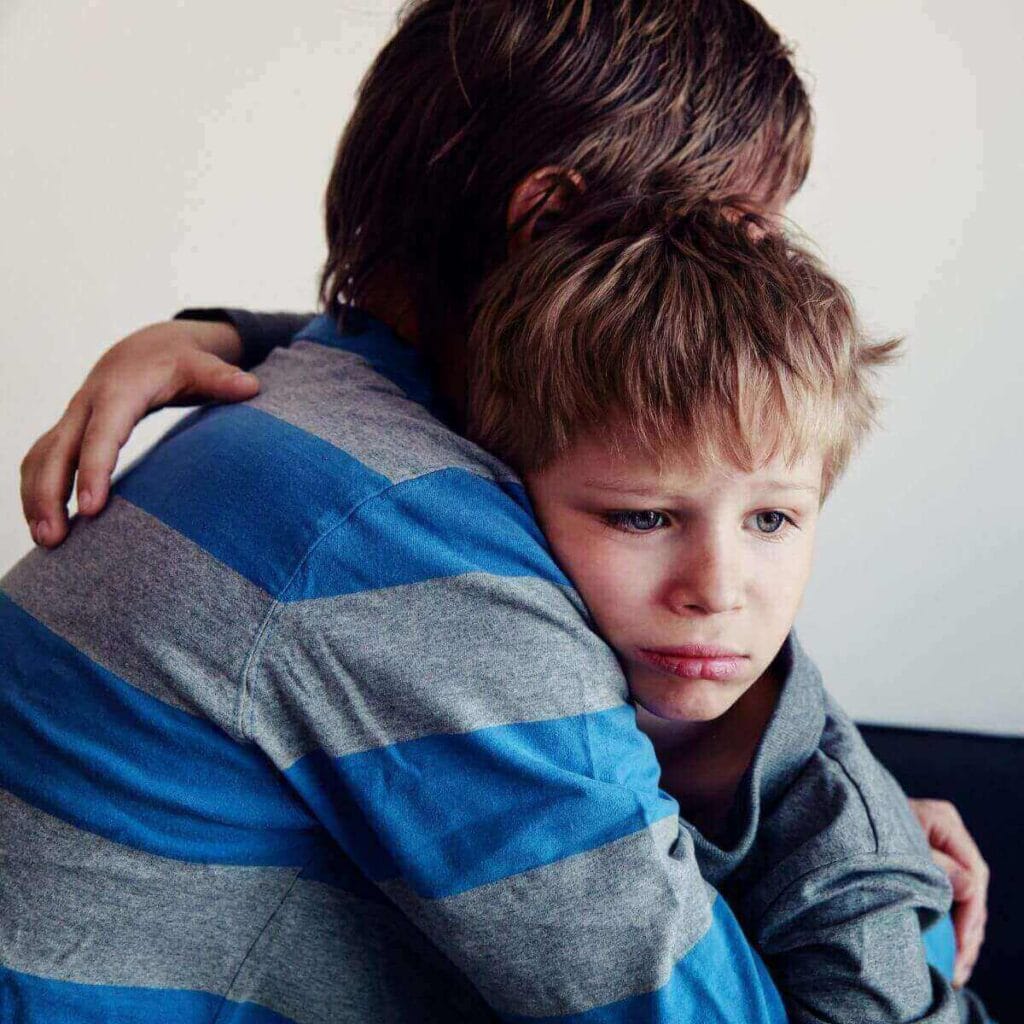 A man wearing a striped long sleeve shirt is hugging a boy wearing a dark grey shirt who looks sad.