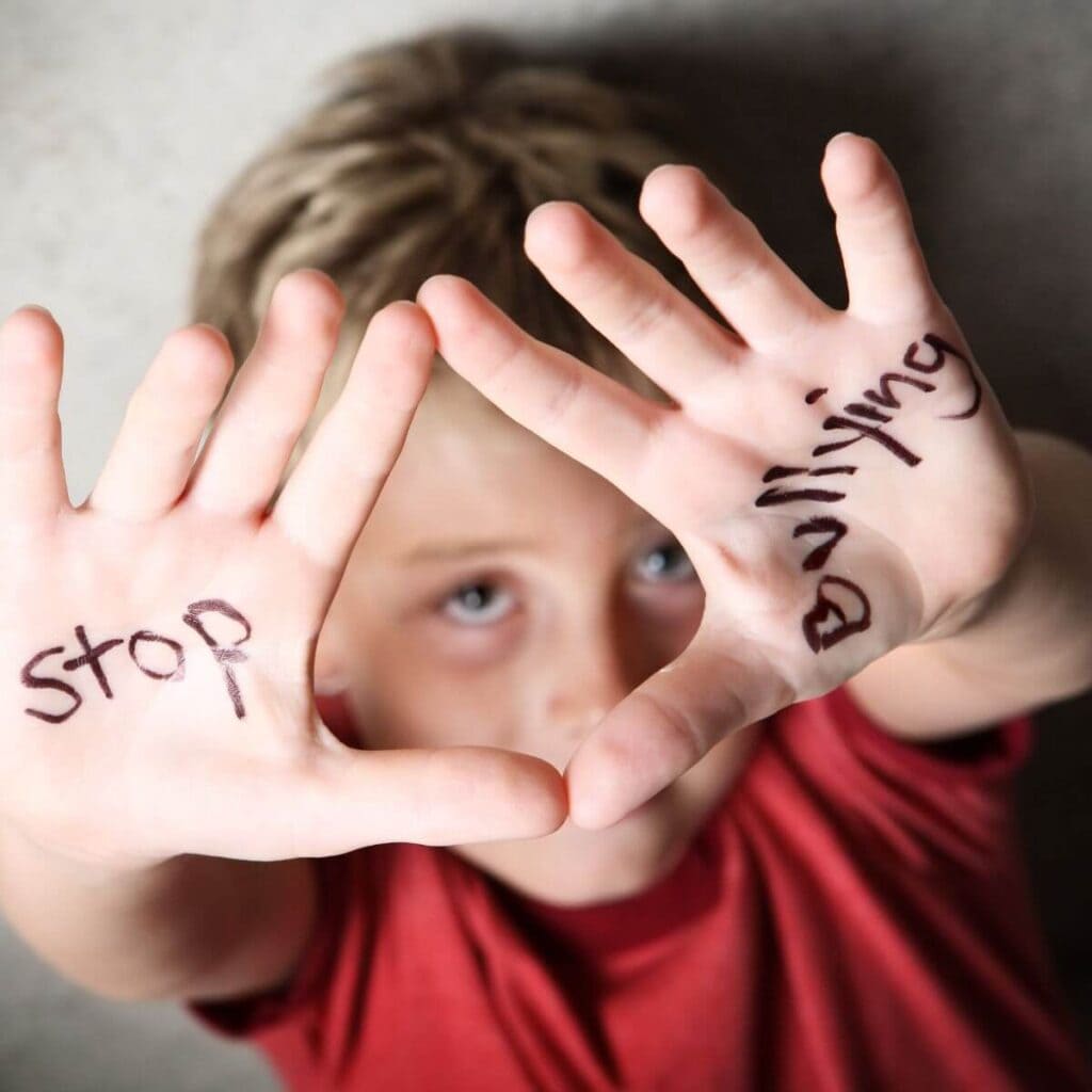 A boy with short dirty blonde hair wearing a red tee-shirt is holding his hands up above his head. On his right plam it says "Stop" and on his left plam it says "Bullying"