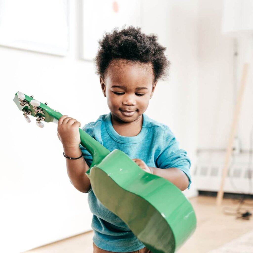 An African American boy wearing a bright blue shirt is standing in the middle of a room with wood floors. He is holding a green guitar.