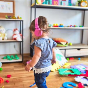 A little girl wearing a blue and white shirt with blue jeans is standing in the middle of a playroom with toys everywhere. She is wearing pink headphones and is smiling.