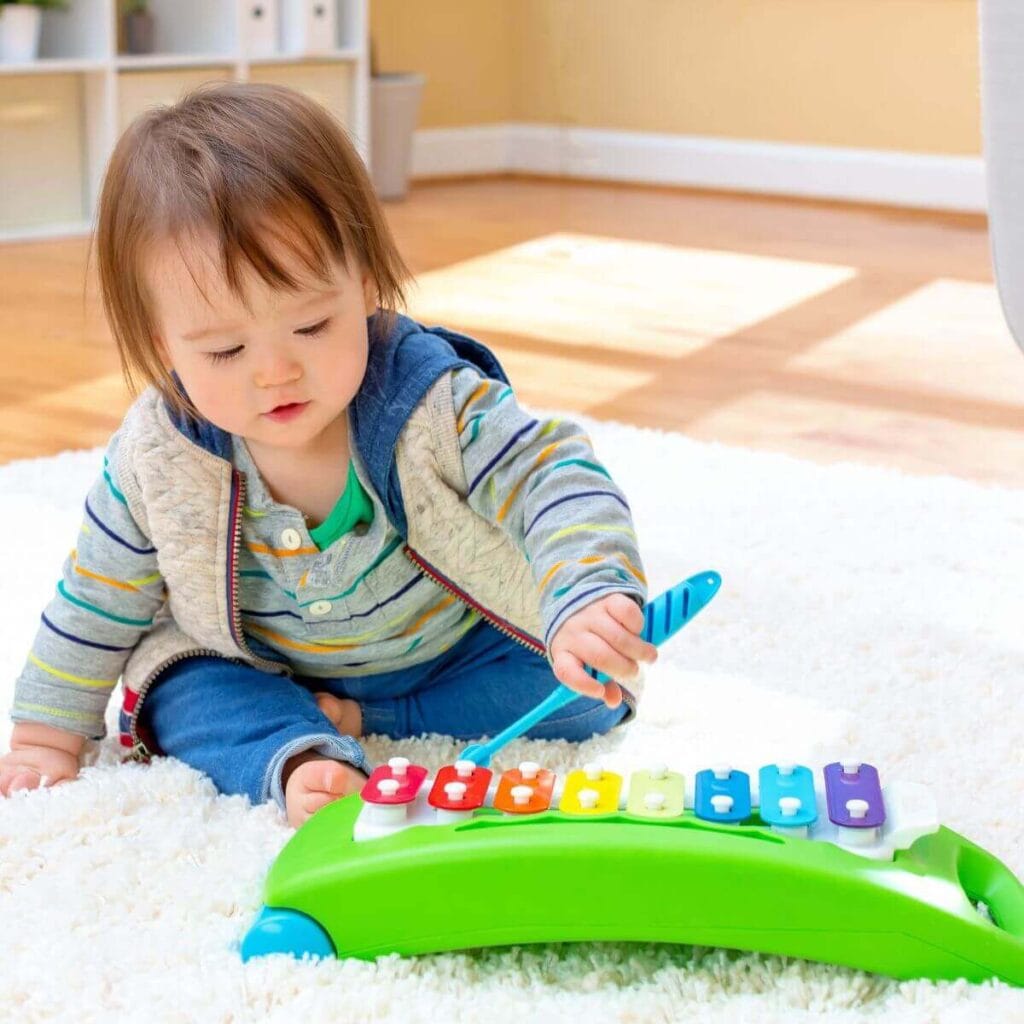 A little one wearing a grey shirt with yellow, blue, green, and orange stripes and blue pants is sitting on a white shag rug playing with a colorful xylophone.