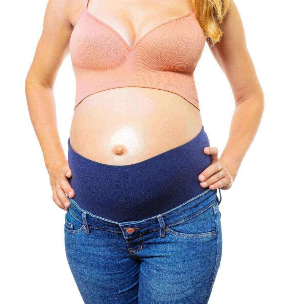 A pregnant woman wearing a rose colored halter shirt with medium blue pregnancy jeans is standing with her hands on her hips.