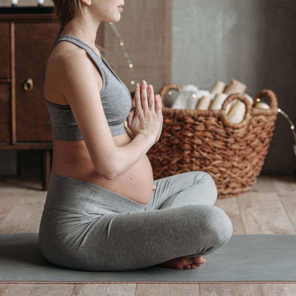 A pregnant woman is sitting cross legged on a yoga mat. She is wearing a grey sports bra and grey stretch pants.