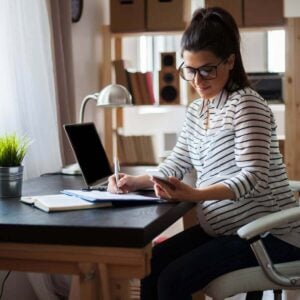 A woman with long brown hair is sitting at a desk. She's wearing black glasses, a black and white striped shirt and black pants. She's writing something down while looking at her phone.