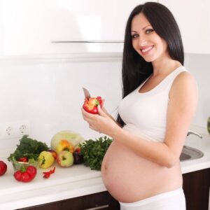 A pregnant woman with long black hair wearing a white tank top and white pants is standing in a kitchen cutting up vegetables.