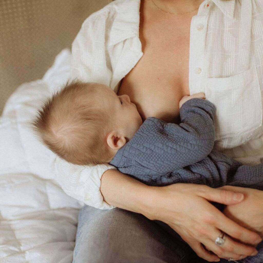 A woman in a white button down shirt is sitting in a chair breastfeeding a newborn baby that is in a dark grey sleeper.