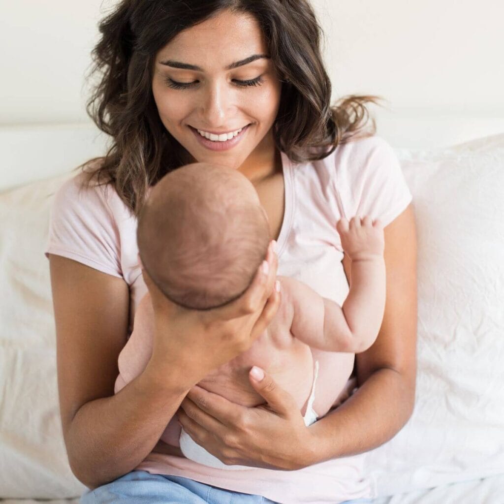 A woman is sitting on a bed with white sheets and a white comforter. She is wearing a light pink tee shirt with light blue jeans. She is smiling down at the baby in her hands that is wearing just a diaper.