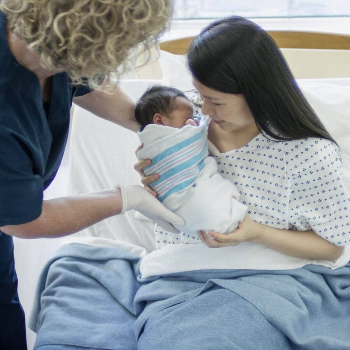 An Asian American woman with long black hair is sitting in a hospital bed. A nurse with blonde curly hair, wearing white gloves and dark blue scrubs is handing a newborn baby to the woman.
