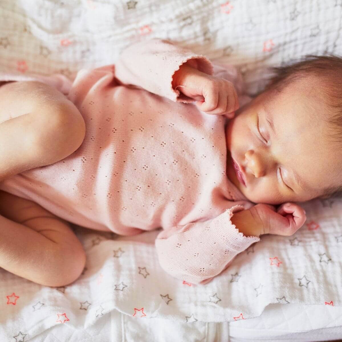 A newborn baby girl is laying on a blanket with stars on it. She is wearing a long sleeve light pink onesie with flowers cut into the fabric and she is sleeping.
