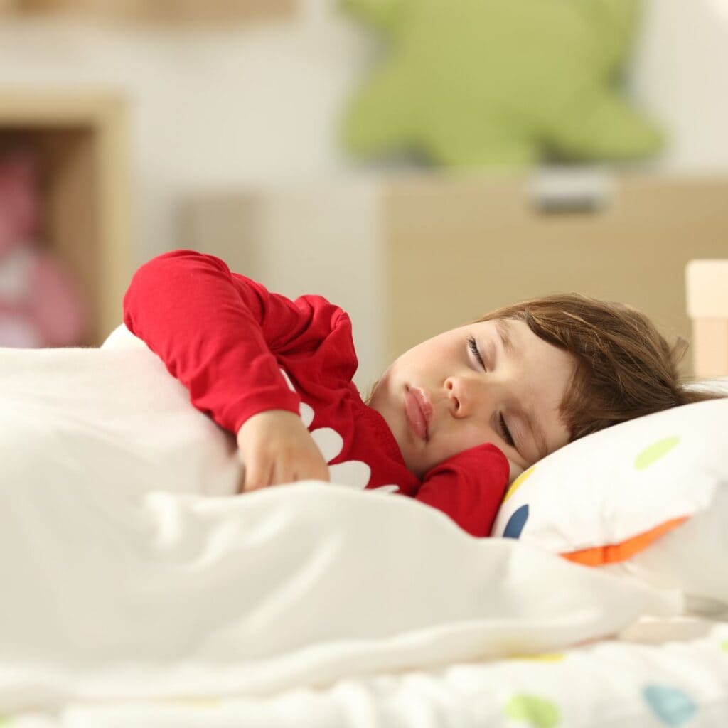 A boy with short brown hair wearing a red long sleeve shirt is laying down in a bed with a white sheet overtop of him. His eyes are closed.