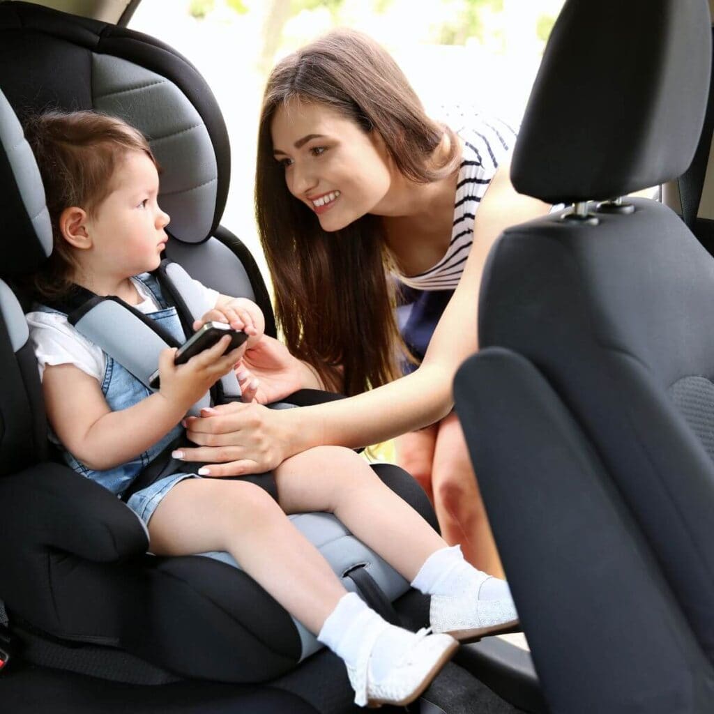 A woman with long brown hair is smiling at a toddler that is sitting in a car seat while she buckles her in.