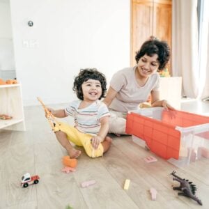 A woman with short black hair is sitting on a wooden floor with a clear plastic bin in front of her with a few toys in it. There is a toddler boy sitting beside her with a giraffee in each hand and he is smiling.