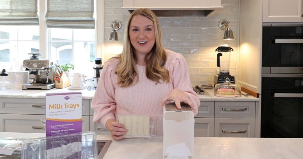 a blonde woman in a pink shirt stands in a kitchen, enthusiastically showing off two different breastmilk storage solutions. Her left hand is on top of the Milkies milk bag freezer storage organizer, while her right hand is holding Milkies Milk Trays with 5 sticks of frozen breastmilk in it.