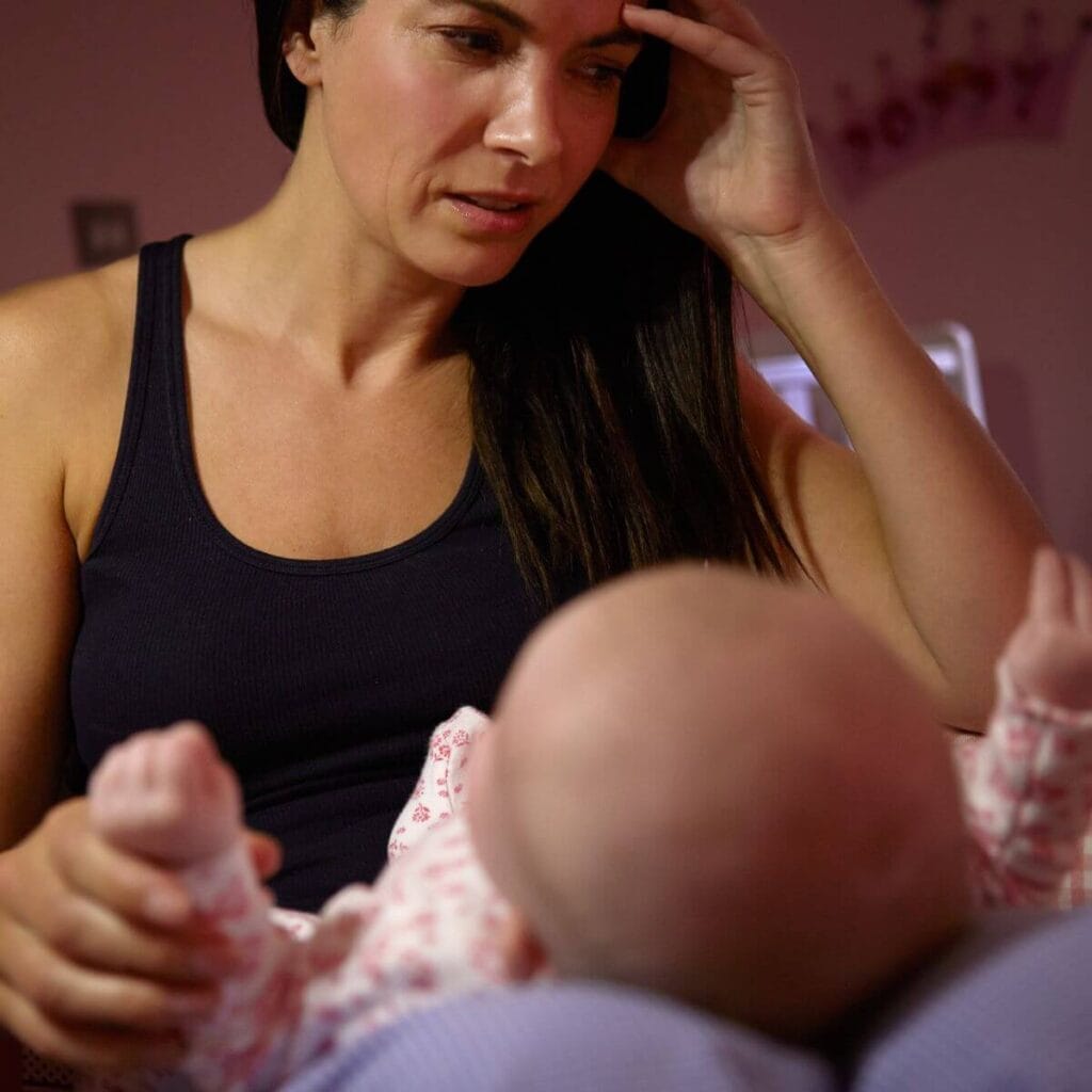 A woman in a black tank top is sitting on a bed. She's got one hand against the side of her face and she's got a baby laying in her lap.