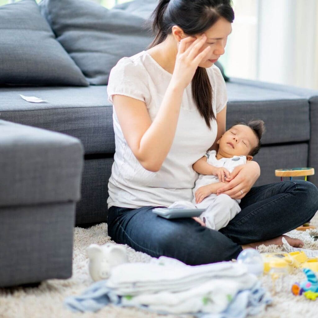 An Asian American woman with dark blue jeans and a white shirt is sitting on the floor in front of a blue denim couch. She is holding a baby in her left arm and she has her right hand up against the side of her head.