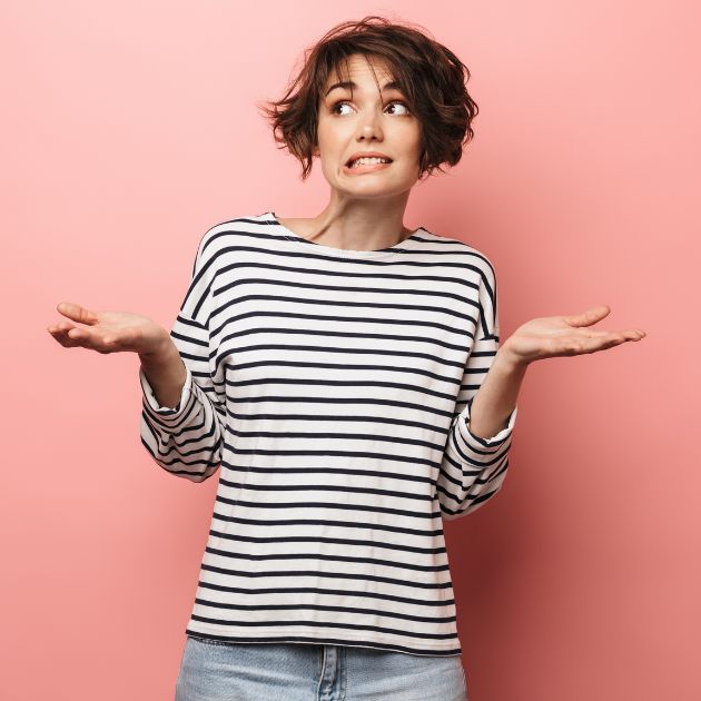 A woman is standing in front of a medium pink background. She is wearing light colored jeans and a white and black striped shirt. She has both hands with palms facing up with a questioning look on her face.