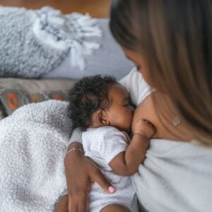 An African American woman is wearing a cream-colored long-sleeve shirt. She is sitting on a couch with a newborn in her arms that is breastfeeding.