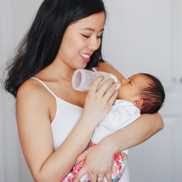 A woman with medium-length black hair, wearing a white spaghetti strap top is standing in front of a white wall. She's holding a newborn while bottle feeding her.