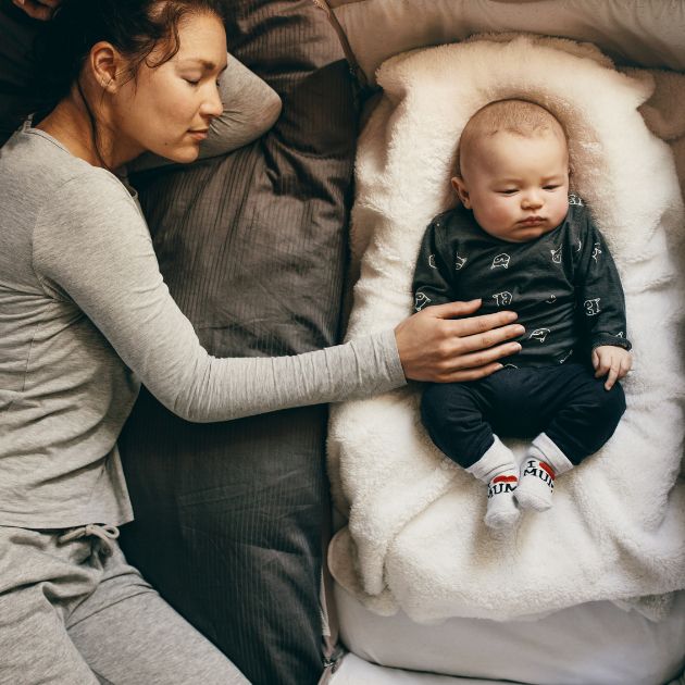 A woman in light grey pajamas is laying on a bed sleeping. A baby is laying next to her and she has her hand on his belly.