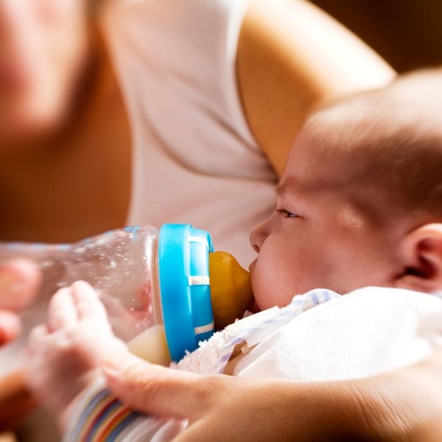 A baby is laying in a woman's arms drinking from a clear bottle with a blue and white striped top on it.