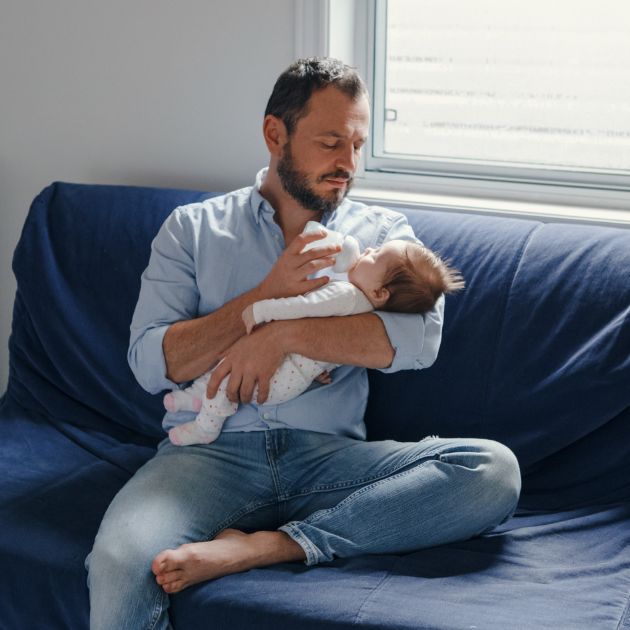 A man in a light blue shirt and jeans is sitting on a dark blue couch. He is holding a newborn while bottle feeding.