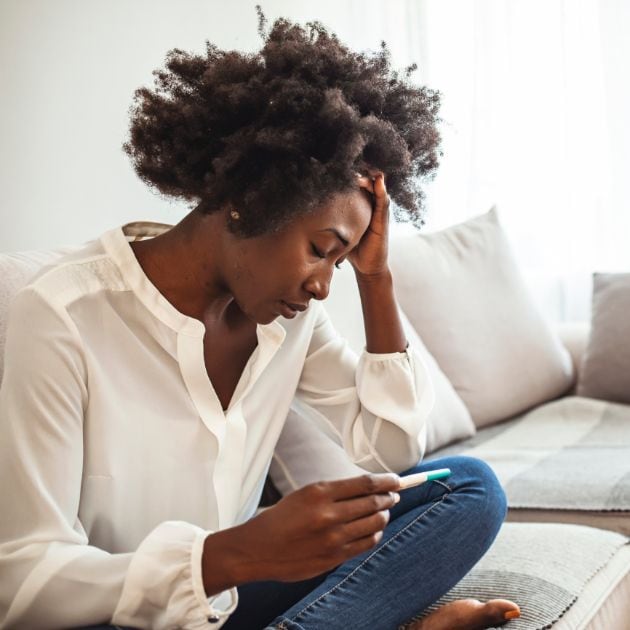 An African American woman is sitting on a couch with her legs up. She is holding a pregnancy test in her hand while looking down with her hand on her head.