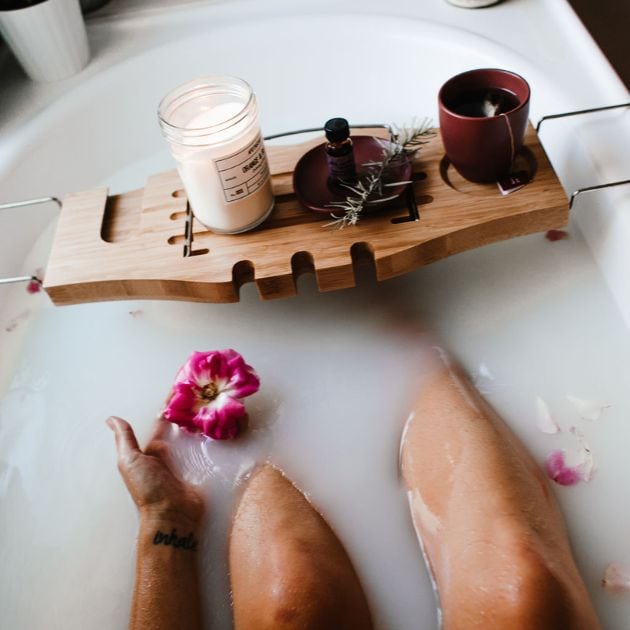 A woman is sitting in a milk bath. She has a wooden board across the tub that has a candle, essential oils, and a cup of tea on it.