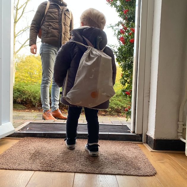 A boy with a white and grey bookbag is walking our of the front door of a house towards his dad who is waiting for him.