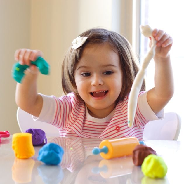 A girl with a pink and white striped shirt is smiling and sitting in front of a white table. On the table is a yellow and blue plastic rolling pin and purple, yellow, blue, green, white, and brown PlayDoh.