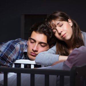 A man and woman are leaning against the side of a crib with their eyes closed.