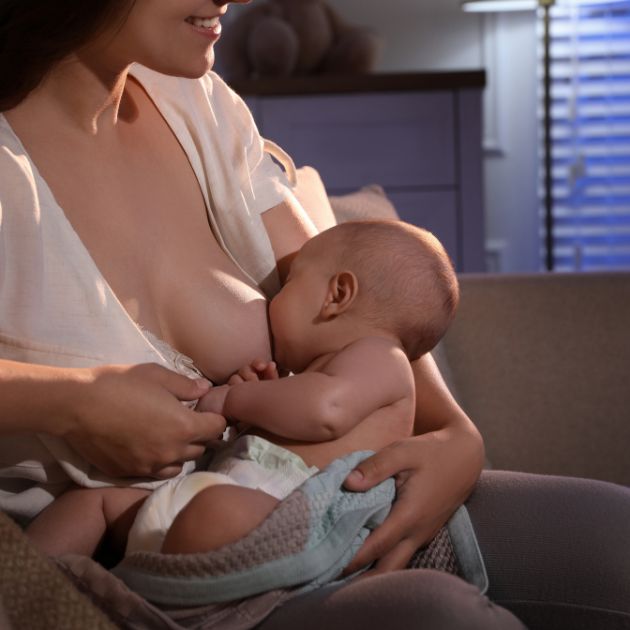 A woman is sitting in a chair in a nursery breastfeeding a baby at nighttime.