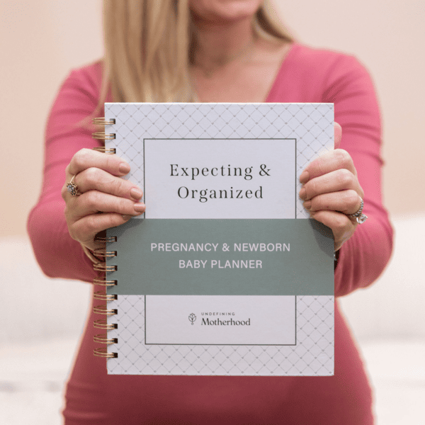 A woman in a vibrant pink dress holds a gold spiral-bound book with a white and sage green cover entitled Expecting & Organized Pregnancy & Newborn Baby Planner out in front of her with both hands