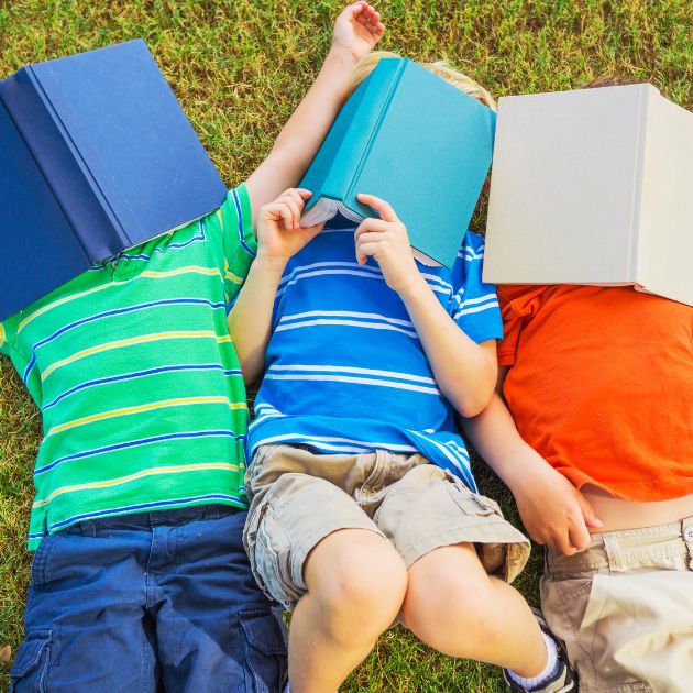 Three boys are laying on their backs in the grass. One is wearing a green striped shirt with a blue book over his face. One is wearing a blue and white striped shirt with a teal book over his face. The last boy is wearing an orange shirt with a cream colored book over his face.