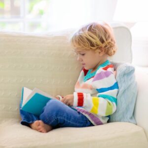 A girl wearing a red, white, yellow, green, teal, blue and purple striped sweater is sitting on a cream colored couch with a blue book open in her lap.