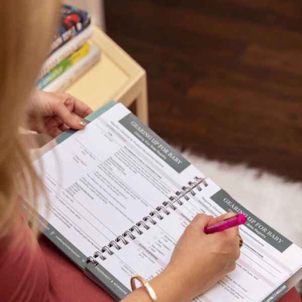 A pregnant woman in a bright pink dress sits with the Expecting and Organized pregnancy planner open on her lap while holding a bright pink pen in her right hand and about to take notes on a page of the pregnancy planner book that reads "gearing up for baby."