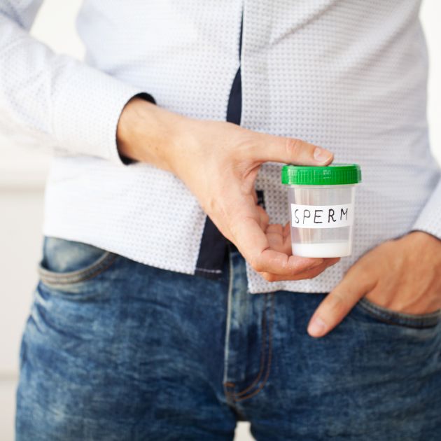 A man in a button-down white shirt and blue jeans is holding a clear cup with a green top that says "Sperm" on the front of it.