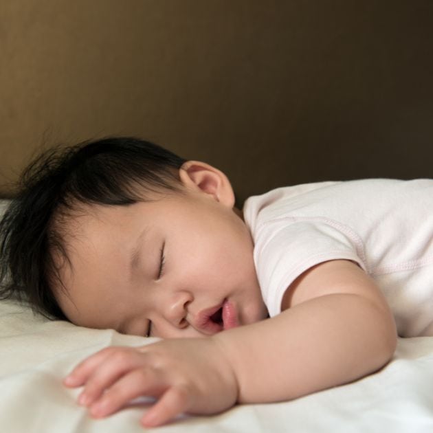 An Asian American baby boy is sleeping on his stomach on a white blanket with a white short sleeve onesie on.