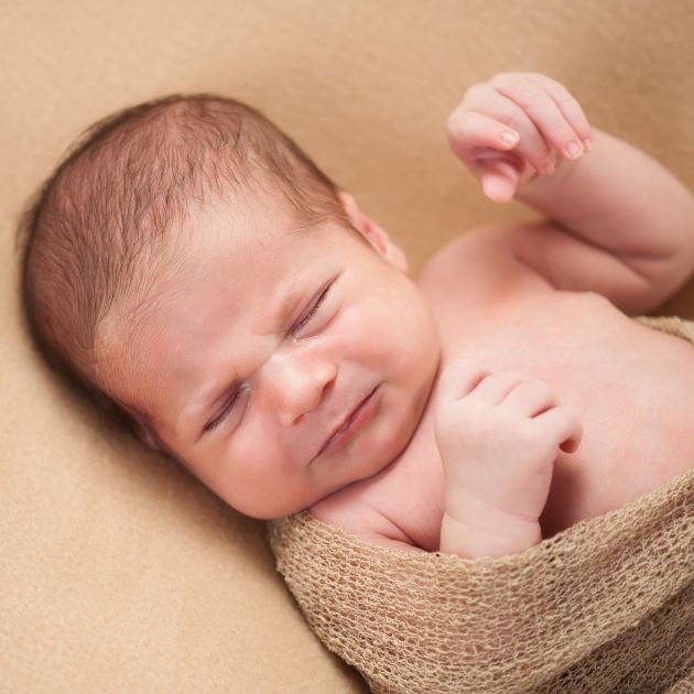 A baby with eyes closed and their face scrunched up in a fussy manner is lying on a cream colored blanket.